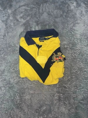 Polo Ralph Lauren County Riders Jockey Club Polo - sportig pikétröja  från Polo Ralph Lauren med korta ärmar. Tröjan har en slående gul grundfärg med kontrasterande mörkblå detaljer på krage, ärmslut och ett stort V-format mönster över bröstet. Den är prydd med en broderad logotyp för "County Riders & Jockey Club" på bröstet och ett tryck på ärmen. En unik och uttrycksfull tröja.  • Skick: Begagnat skick med patina. Det finns några fläckar och mindre hål – vänligen se de sista bilderna för en tydlig bild av skicket.