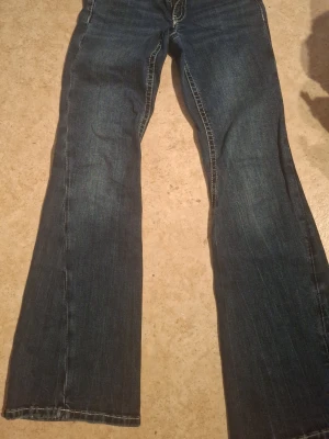 Mörkblå bootcut jeans - Säljer ett par mörkblå bootcut jeans med klassisk femficksdesign och kontrastsömmar. Jeansen har utsvängda ben med snygg tvätt. Perfekta för dig som gillar retrostil och vill ha ett par bekväma jeans med cool passform.