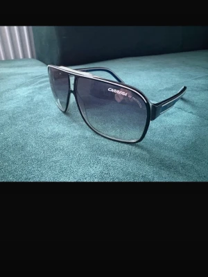 Carrera solglasögon  - Snygga pilotsolglasögon från Carrera med svart front och blå detaljer på skalmarna. Glasen är tonade i grått och bågarna har en sportig, retroinspirerad design. Perfekta för dig som vill sticka ut med en cool accessoar.