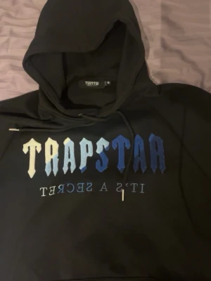 Trapstar hoodie med blått tryck - Säljer en svart hoodie från Trapstar med stor huva och dragsko. På bröstet står det 'TRAPSTAR' i gradient från vitt till blått och texten 'IT'S A SECRET' spegelvänt under. Klassisk känguruficka framtill och ribbade muddar. Perfekt för streetwear-stilen.