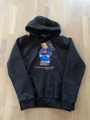Svart hoodie Polo Bear Ralph Lauren - Svart hoodie från Polo Ralph Lauren med ikoniska Polo Bear-trycket på bröstet. Klassisk huva och stor magficka framtill. Tillverkad i mjukt material som känns skönt mot huden. Perfekt för dig som vill ha en stilren och trendig look.