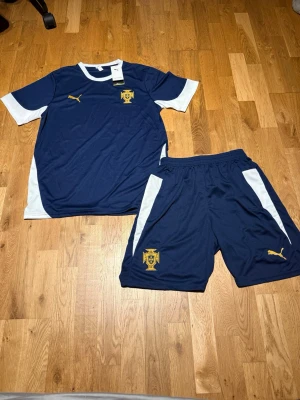 Portugal summer set Navy blue  - Snyggt blått fotbollsställ från Puma med matchande tröja och shorts. Setet har vita detaljer på ärmar och sidor samt gula Puma-loggor och klubbmärke på både tröja och shorts. Tillverkat i lätt och funktionellt material som passar perfekt till träning eller match.