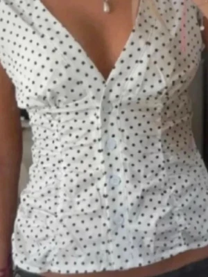 Vit prickig blus med v-ringning - Söt vit blus med svarta prickar och djup v-ringning. Toppen har knappar framtill och figurnära passform. Passar som S.