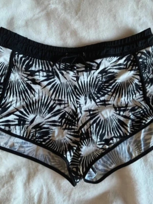Svarta och vita badbyxor med palmtryck - Säljer ett par snygga badshorts i svart och vitt med tropiskt mönster. Shortsen har elastisk midja med snörning framtill och svarta kanter. Perfekta för stranden eller poolen. Materialet känns som syntet, snabbtorkande och bekvämt. 