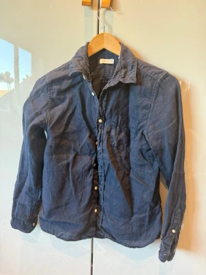 Mörkblå linneskjorta från Gant - Snygg mörkblå skjorta från Gant i linne med klassisk krage och vita knappar framtill. Skjortan har lång ärm och en bröstficka, perfekt för en avslappnad men ändå stilren look.