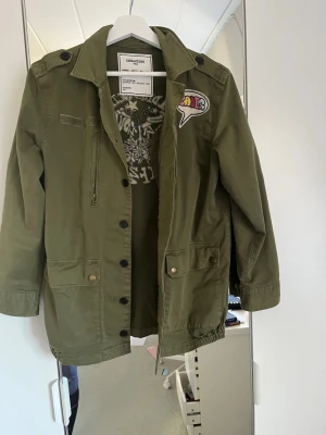 Olivgrön vårjacka från Zadig & Voltaire - Snygg olivgrön vårjacka från Zadig & Voltaire med coola broderade detaljer och patch på bröstet. Jackan har flera fickor, knappstängning och en unik ryggdesign med färgglada broderier och text. Perfekt för dig som vill sticka ut med din stil.