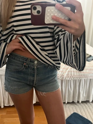 Levi's blå jeansshorts - Säljer ett par klassiska blå jeansshorts från Levi's med hög midja och råa kanter. Shortsen har fem fickor, knappgylf och en avslappnad passform. Perfekta till sommaren och lätta att matcha med olika toppar.