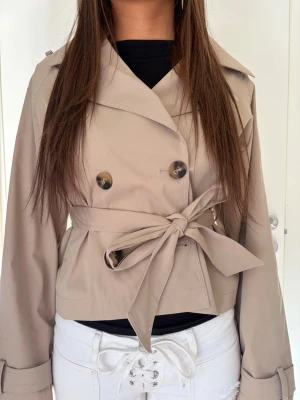 Kort beige trenchcoat med knytskärp - Beige trenchcoat i kort modell med dubbelknäppning och stora knappar framtill. Jackan har ett brett knytskärp i midjan och klassiska axelklaffar. Från Gina tricot 14+ i storlek S 