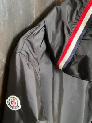 Svart Moncler vindjacka med huva - Säljer en stilren Moncler vindbreaker i mycket bra skick. Jackan är lätt, bekväm och perfekt för både vardag och vår/sommar.  Utsidan är i toppskick utan några synliga tecken på användning. Insidan är något blekt i materialet, men det är inget som märks när man bär jackan.