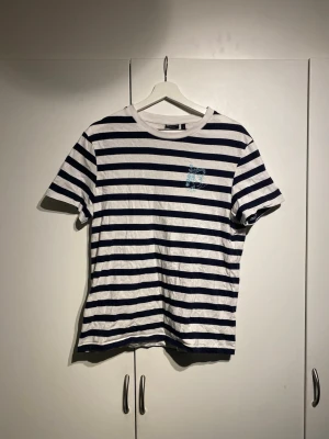 Randig t-shirt från Asos - Säljer en vit och marinblå randig t-shirt från Asos med korta ärmar. T-shirten har en liten broderad detalj på bröstet och är gjord i mjuk bomull. Perfekt för en avslappnad och stilren look.
