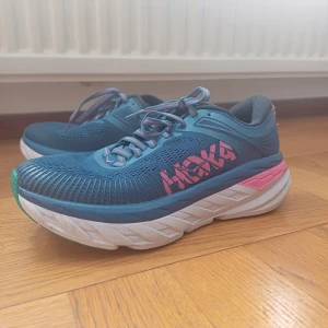 Blå Hoka sneakers med rosa detaljer - Snygga blå sneakers från Hoka med rosa och vita detaljer samt tjock vit sula. Ovandelen är i mesh för bra ventilation och skorna har klassisk snörning. Perfekta för dig som gillar sportig stil och vill ha bekväma skor med färgglad touch.