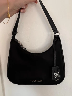 Svart axelremsväska Steve Madden - Snygg svart axelremsväska från Steve Madden i en stilren, rektangulär form. Väskan har silverfärgade detaljer, dragkedja upptill och ett dekorativt SM-tag. Tillverkad i ett slätt, tåligt material som ger en modern look.