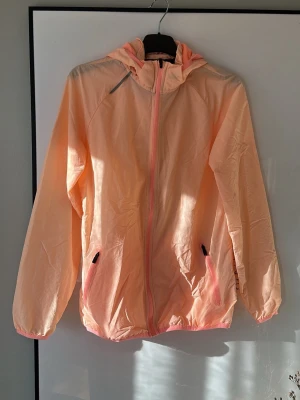 Persikofärgad vindjacka med huva - Snygg persikofärgad vindjacka från SOC med huva och dragkedja framtill. Jackan har två fickor med dragkedja, elastiska muddar och reflekterande detaljer på axel och rygg. Tillverkad i lätt och tunt material, perfekt för blåsiga dagar. Pris kan diskuteras.