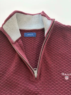 Vinröd half zip tröja från GANT - Snygg vinröd tröja från GANT med strukturstickat mönster och halv dragkedja. Tröjan har hög krage med kontrastfärgad insida och broderad logga på bröstet. Perfekt till både jeans och chinos för en clean look.