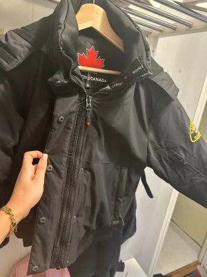 Svart parkas från Canada zavetti  - Snygg svart parkas från Canada Goose med klassisk patch på ärmen och stor huva. Jackan har dragkedja och knappar framtill, flera fickor och justerbar midja. Perfekt för kalla dagar och riktigt varm. Materialet är slitstarkt och vindtätt.