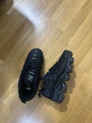 Nike Air Vapormax Plus svarta sneakers - Svarta Nike Air Vapormax Plus sneakers med futuristisk design och synliga luftbubblor i sulan. Ovandelen är i mesh och syntet med svarta detaljer och vita accenter på tån. Skorna har snörning och en chunky sula som ger en sportig vibe.