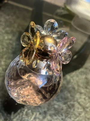 Parfymflaska med blommig kristallkork - Unik parfymflaska i facetterat glas med en dekorativ kork formad som en blomma i transparent och ljuslila kristall. Flaskan har en rund, elegant form och detaljer i guld. Perfekt som inredningsdetalj eller för din favoritdoft.