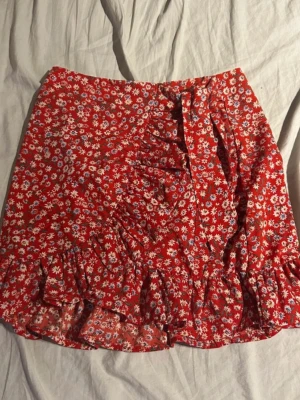 Röd blommig kjol - Supersöt röd kortkjol med vitt blommönster och volangkant. Omlottdesign med knytband i sidan som ger en fin och feminin passform. Perfekt till sommaren eller semestern matcha med linne och sandaler för en fräsch look.(ifrån shein)