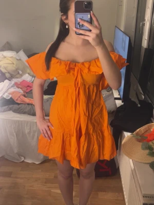Orange offshoulder kortklänning - Snygg orange kortklänning med offshoulder och volangdetaljer. Klänningen har en söt knytning framtill och är tillverkad i ett lätt tyg, perfekt för varma dagar. Passformen är loose och modellen är luftig och bekväm.