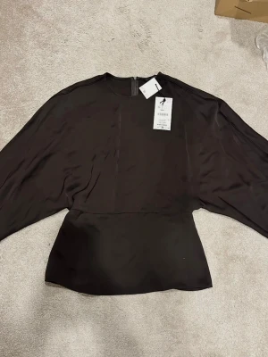 Svart blus med vida ärmar från Zara - Snygg svart blus från Zara med rund hals och vida långärmade ärmar. Blusen har en slät, glansig yta och markerad midja som ger en cool siluett. Perfekt för att skapa en trendig look.