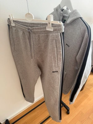 Grå mjukisbyxor från BOSS - Snygga grå mjukisbyxor från BOSS med svarta sidoremsor och dragsko i midjan. Byxorna har två fickor fram och en avslappnad passform. Perfekta för chill dagar eller när du vill ha en sportig look.
