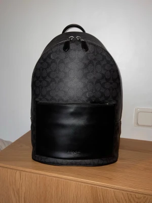 Coach Metropolitan ryggsäck - Snygg svart ryggsäck från Coach med klassiskt monogrammönster och stor slät läderficka framtill. Ryggsäcken har dubbla dragkedjor upptill, bärhandtag och justerbara axelband. Perfekt för dig som vill ha en stilren och praktisk väska. Använd väldigt få gånger.