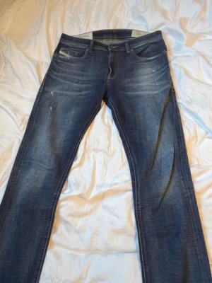 Blå jeans från Diesel med slitningar - Säljer ett par snygga blå jeans från Diesel med klassisk femficksmodell och coola slitningar. Jeansen har en något tvättad look och kontrastsömmar. Märkeslappen syns tydligt på myntfickan. Perfekta för en avslappnad stil.