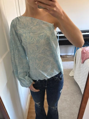 Ljusblå oneshoulder topp med mönster - Säljer en ljusblå oneshoulder topp med vitt blommigt mönster. Toppen har en lång puffärm och är axelbandslös på ena sidan. Perfekt för dig som vill ha något unikt och trendigt till sommaren.