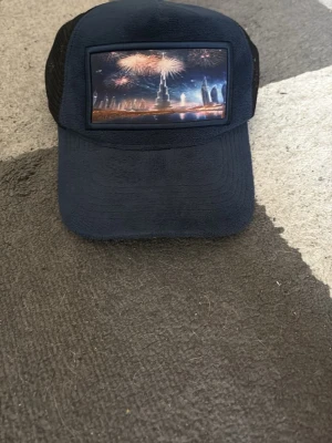 Svart keps med Dubai-motiv - Cool svart keps med mesh baktill och justerbar snapback. Framtill finns en färgglad bild av Dubais skyline och fyrverkerier. Perfekt accessoar för dig som gillar unika motiv och streetstyle.