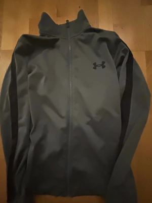 Grå träningsjacka från Under Armour - Snygg grå träningsjacka från Under Armour med hel dragkedja och hög krage. Jackan har svarta detaljer längs ärmarna och en diskret logga på bröstet. Tillverkad i ett mjukt och stretchigt material som passar perfekt till träning eller uppvärmning.
