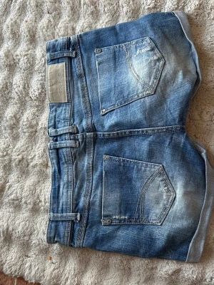 Blå denimshorts med slitningar - Säljer ett par blå denimshorts med slitna detaljer och uppvikta benslut. Shortsen har fem fickor, bälteshällor och dubbla knappar fram. Perfekta för en avslappnad och trendig look.