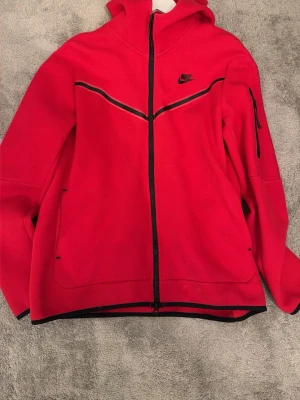 Röd Nike hoodie med dragkedja - Snygg röd hoodie från Nike med hel dragkedja framtill och svarta detaljer. Har huva, fickor på sidorna och en extra ficka med dragkedja på ärmen. Tillverkad i mjukt och stretchigt material, perfekt för en sportig och avslappnad stil. Helt fläck fri 