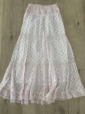 Vit maxikjol med rosa mönster - Lång, luftig maxikjol i vitt med små rosa mönster och bred smock i midjan. Kjolen har volanger och spetsdetaljer som ger en romantisk känsla. Perfekt för sommardagar och gjord i ett tunt, mjukt tyg som känns skönt mot huden.