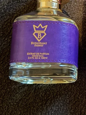 Bujairami Executor Extrait de Parfum - Lyxig parfymflaska från Bujairami Sydney med en unik design. Flaskan är genomskinlig med ett lila band i läder runtom och gulddetaljer på korken. Rymmer 100 ml och har en exklusiv känsla med guldfärgad logotyp och text på framsidan.