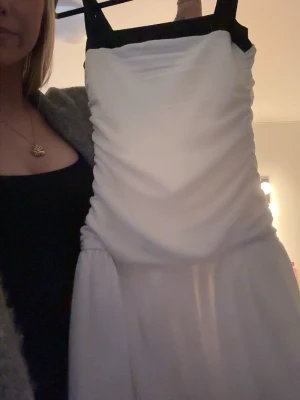 Vit kort klänning med svarta band - Säljer en söt vit kortklänning med svarta axelband. Klänningen har en figurnära passform med lätt rynkad effekt framtill och volang nedtill. Perfekt för dig som vill ha en stilren men ändå unik look.
