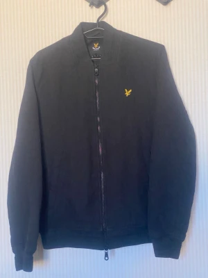 Svart bomberjacka från Lyle & Scott - Snygg svart bomberjacka från Lyle & Scott med klassisk gul logga på bröstet. Jackan har dragkedja framtill, ribbade muddar och krage. Enkel och stilren design som passar till det mesta. Materialet känns mjukt och bekvämt.