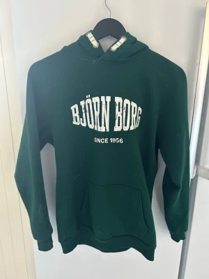 Grön Björn Borg hoodie med tryck - Mörkgrön hoodie från Björn Borg med vit text 'BJÖRN BORG SINCE 1956' på bröstet. Klassisk känguruficka framtill och huva med vitmönstrat foder. Perfekt för en avslappnad och sportig stil. Tillverkad i mjukt bomullsmaterial.