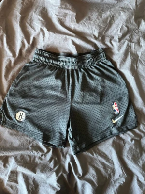 NBA Nets shorts - Snygga, grå, korta shorts från Nike med Brooklyn Nets och NBA-logga på framsidan. Shortsen har elastisk midja, utan snöre och är perfekta för basket eller träning. Luftigt material som ger skön känsla och rörelsefrihet. Shortsen är köpte i NBA affären i New York och jag säljer dem nu då jag inte längre använder dem. De är storlek M för barn, men skulle säga S i normal storlek. 