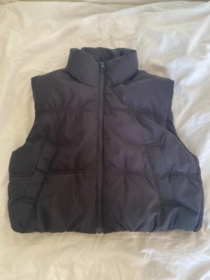 Svart dunväst med hög krage - Svart dunväst med hög krage och dragkedja framtill. Västen har två snedställda fickor. Perfekt att slänga över en hoodie eller tröja för extra värme. Materialet är glansigt och känns mjukt. Inga defekter