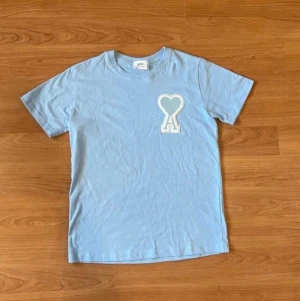 Ljusblå Ami Paris t-shirt med hjärtdetalj - Säljer en ljusblå t-shirt från Ami Paris med korta ärmar och en stor broderad patch på bröstet i form av ett hjärta och bokstaven A. Tillverkad i mjuk bomull och har en relaxed passform. Perfekt för dig som gillar stilrena plagg med en twist. PRIS KAN DISKUTERAS