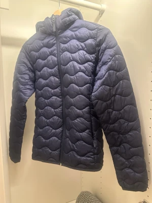 Mörkblå pufferjacka från McKinley - Säljer en mörkblå pufferjacka från McKinley med vågformat quiltat mönster och huva. Jackan har dragkedja framtill och är fodrad med ljusrosa insida. Perfekt för kalla vinterdagar och riktigt skön att bära.