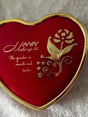 Hjärtformad Makeup Set  - Söt hjärtformad ögonskuggspalett från MINYN med spegel. Innehåller nio olika färger i hjärtformade pans: vit, gul, grön, svart, blå, rosa, orange, persika och lila. Asken är röd med gulddetaljer och blommotiv. Perfekt för dig som gillar färgglada och kreativa sminkningar.