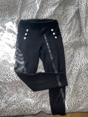 Svarta leggings med knappar från SHEIN - Säljer ett par svarta leggings från SHEIN med hög midja och dekorativa silverfärgade knappar framtill. Stretchigt material som sitter skönt och formar sig efter kroppen. Perfekta till en snygg outfit när du vill ha något bekvämt men ändå stilrent.