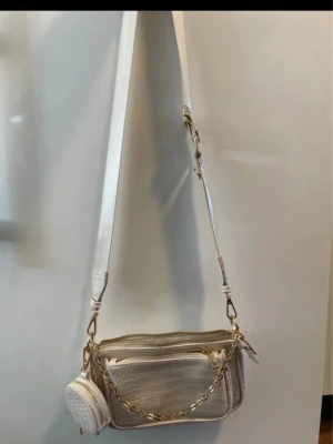 Beige axelväska med kedjedetalj - Snygg beige axelväska med croco-mönster och guldfärgad kedja framtill. Väskan har justerbar axelrem och ett litet rundat fack på sidan. Perfekt för dig som gillar trendiga accessoarer och stilren design.