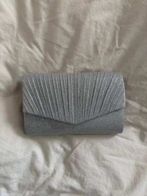 Silverfärgad glittrig balväska - Snygg silverfärgad balväska med glittrigt tyg och veckad framsida. Väskan har en klassisk rektangulär form och stängs med ett lock. Perfekt accessoar för att lyfta din outfit med lite extra bling.