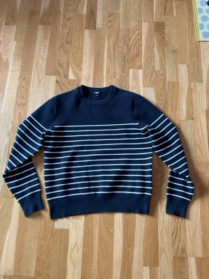 Mörkblå randig tröja från Uniqlo - Snygg mörkblå tröja från Uniqlo med vita horisontella ränder över bröstet och ärmarna. Tröjan har rund halsringning och långa ärmar. Perfekt för en stilren och avslappnad look.