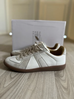 Vita sneakers från Maison Margiela - Snygga vita sneakers från Maison Margiela med beige detaljer och bruna gummisulor. Skorna har en klassisk låg modell med paneler i läder och mocka, samt matchande beige snörning. Perfekt för dig som gillar stilrena och trendiga sneakers.
