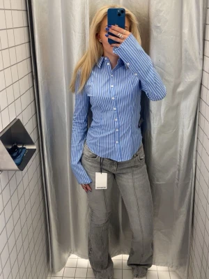 Blårandig skjorta från Bershka  - Snygg blå skjorta med vita vertikala ränder. Skjortan har klassisk krage, långa ärmar och knäppning framtill. Figurnära passform och tillverkad i ett mjukt bomullsmaterial som känns bekvämt hela dagen. Den är figursydd och har utsvävningar vid underarmen.