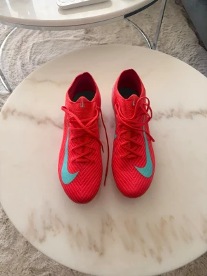 Nike Mercurial elite - Säljer ett par röda Nike Mercurial Elite FG med turkosblå swoosh. Skorna har snörning och är tillverkade i ett lätt syntetmaterial som ger bra bollkontroll och passform. Perfekta för dig som vill sticka ut på planen med både stil och funktion. Endast används under ett inhopp på 35 min.