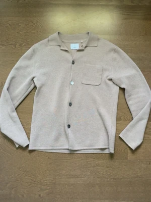 Beige skjorta från Bläck med knappar - Stilren beige skjorta från Bläck med klassisk krage, bröstficka och knäppning framtill. Tröjan är köpt från MQ för 800kr. Skjortan är långärmad och har en mjuk känsla, perfekt för en avslappnad men snygg look.